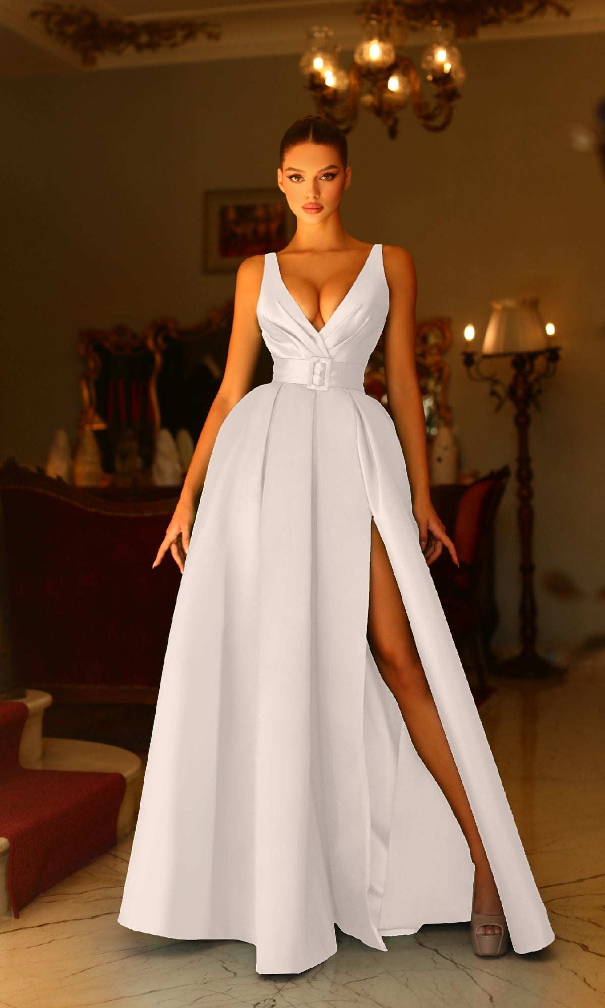 A woman in a Belted Long White Prom Dress—Tina Holly BB209W, featuring a deep V-neckline and high slit—stands in an elegant, dimly lit room with vintage decor.